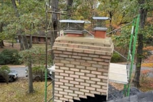 Brick chimney with two metal caps and mesh guards, surrounded by autumn trees and scaffold.